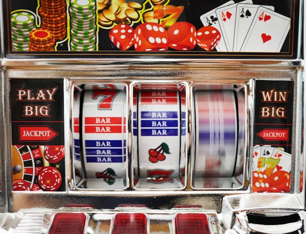 A close-up view of a slot machine with vibrant colors and various gambling symbols such as dice, coins, and playing cards. The reels display a combination of symbols, including bars, cherries, and the number seven. The chrome exterior and colorful details convey the theme of casino and gambling.