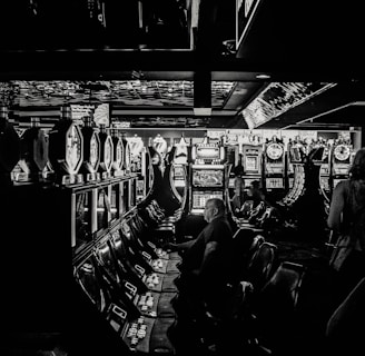 A dimly lit casino filled with rows of slot machines, some occupied by people. The machines' lights contrast with the dark surroundings, creating a vibrant yet subdued atmosphere.