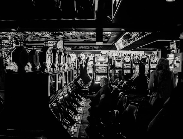 A dimly lit casino filled with rows of slot machines, some occupied by people. The machines' lights contrast with the dark surroundings, creating a vibrant yet subdued atmosphere.