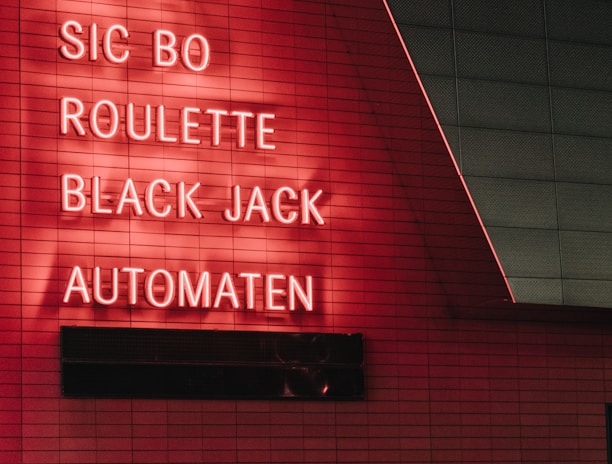 Red neon letters are mounted on a dark tiled wall, displaying the words Poker, Sic Bo, Roulette, Black Jack, and Automaten.