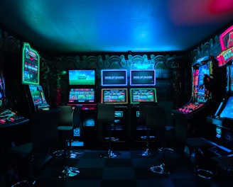 Dimly lit gaming arcade featuring a variety of brightly illuminated slot machines and video gaming screens. Neon signs and colorful displays create a vibrant atmosphere. The arrangement of gaming machines suggests an inviting space for entertainment, with chairs positioned for players.