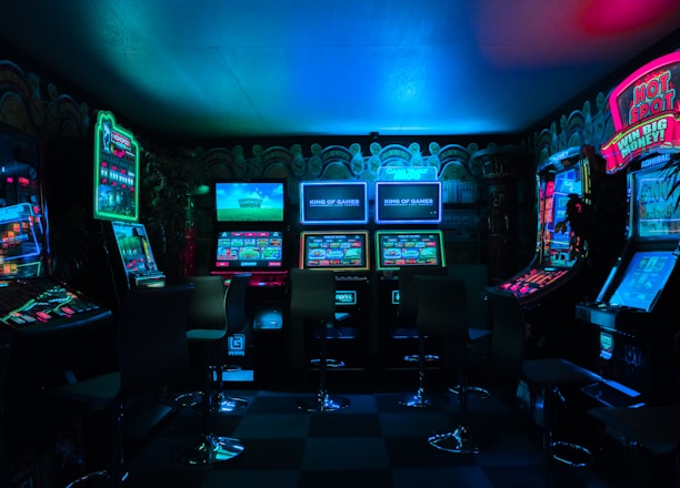 Dimly lit gaming arcade featuring a variety of brightly illuminated slot machines and video gaming screens. Neon signs and colorful displays create a vibrant atmosphere. The arrangement of gaming machines suggests an inviting space for entertainment, with chairs positioned for players.