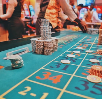A casino scene with a roulette table features prominently, covered in green felt with colorful numbers and betting squares. Several stacks of poker chips are placed on different numbers, and the hands of several people are visible as they interact with the table. In the background, there are slot machines and more people, creating a vibrant and busy atmosphere.