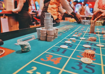A casino scene with a roulette table features prominently, covered in green felt with colorful numbers and betting squares. Several stacks of poker chips are placed on different numbers, and the hands of several people are visible as they interact with the table. In the background, there are slot machines and more people, creating a vibrant and busy atmosphere.