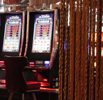 A pair of casino slot machines, each with detailed screens displaying various game outcomes and jackpot amounts. The chairs in front of the slot machines are dark and cushioned for comfort. The background features a hanging curtain made of golden ropes and other casino decorations are visible.