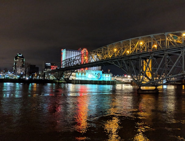 A brightly lit casino with neon signs and red lights is reflected in a river. A bridge stretches across the water, with an urban skyline visible in the background against an overcast night sky.