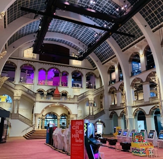 The interior of a large, ornately designed building with arched ceilings and intricate architectural details. The space features gaming machines lined up along the floor, under a high ceiling with a grid pattern. Lighting in purple and white tones enhances the ambiance.