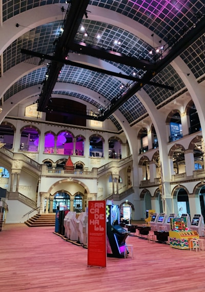 The interior of a large, ornately designed building with arched ceilings and intricate architectural details. The space features gaming machines lined up along the floor, under a high ceiling with a grid pattern. Lighting in purple and white tones enhances the ambiance.