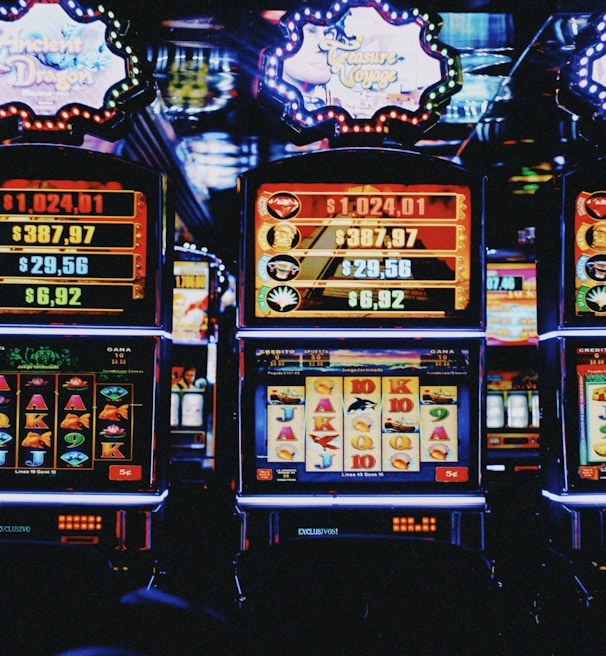 A row of colorful slot machines lit with bright neon lights, showcasing various graphical elements on the screens, indicating different games and jackpots. The atmosphere suggests a lively casino setting, bustling with excitement.