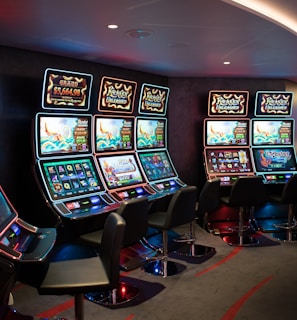 A row of brightly lit slot machines in a casino setting, each with colorful displays and various game themes. The machines are lined up against a dark wall and are unoccupied, with cushioned chairs placed in front of them. The area is dimly lit except for the glow from the screens, creating a vibrant and engaging atmosphere.